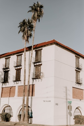 A white building with a Spanish architectural style features red-tiled roofs and multiple balconies with iron railings. Tall palm trees stand in front of the building, and there is minimal shadowing on the walls. The corner of the building shows street signs and is labeled 'The Village' and numbered '1418'.