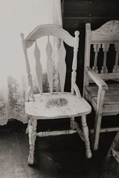 An old, worn wooden chair with intricate backrest designs stands next to a window adorned with lace curtains. The chair has visible signs of age and use, adding a rustic charm to the scene. Next to it is another wooden piece, possibly a chair or part of a bench, featuring decorative carvings.