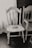 An old, worn wooden chair with intricate backrest designs stands next to a window adorned with lace curtains. The chair has visible signs of age and use, adding a rustic charm to the scene. Next to it is another wooden piece, possibly a chair or part of a bench, featuring decorative carvings.