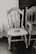 An old, worn wooden chair with intricate backrest designs stands next to a window adorned with lace curtains. The chair has visible signs of age and use, adding a rustic charm to the scene. Next to it is another wooden piece, possibly a chair or part of a bench, featuring decorative carvings.