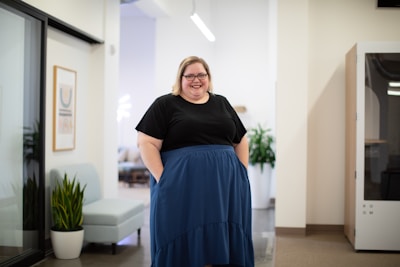A person is standing in a modern office environment, smiling. They are wearing a black top and a blue skirt, and their hands are in their pockets. The office features a light color scheme with a few plants and minimalistic furniture.