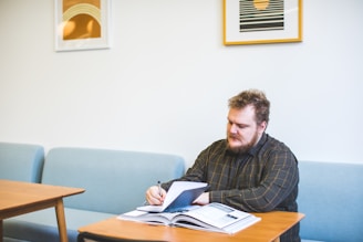 A person with a beard sits at a wooden table writing in a notebook. The setting is a room with light blue couches and framed abstract art on the walls. Natural light brightens the room, creating a calm and focused atmosphere.
