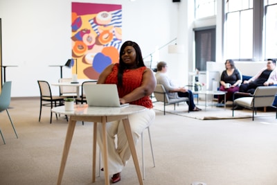 A woman is sitting at a small round table in an open office space, using a laptop. Around her, several people are sitting in a lounge area engaged in conversation. The room is decorated with a large, colorful abstract painting on the wall and has large windows allowing natural light to enter.