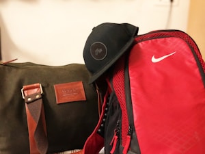 A green duffel bag with brown accents, featuring a leather patch displaying the brand name 'Volcoo', sits alongside a bright red backpack with a prominent white logo. A black cap rests on top of the backpack.