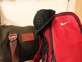 A green duffel bag with brown accents, featuring a leather patch displaying the brand name 'Volcoo', sits alongside a bright red backpack with a prominent white logo. A black cap rests on top of the backpack.