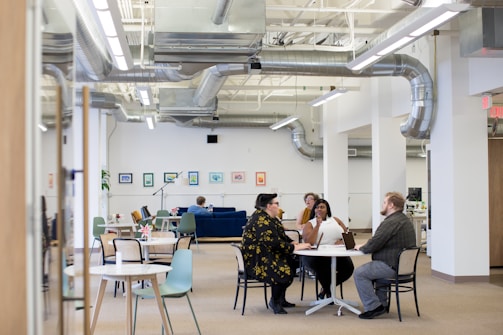 A spacious and modern office with high ceilings and visible ductwork. Four people are gathered at a round table, engaged in discussion. The room features a variety of seating options, including chairs and a couch in the background. Walls are adorned with framed art, and there is a plant in the corner.
