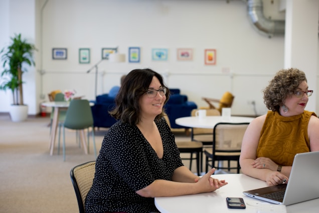 Two people are seated at a round table, engaged in conversation. One person is wearing a polka dot dress and the other is in a mustard-colored top. The room has a casual, cozy atmosphere with several chairs, a plant, and framed art on the walls.