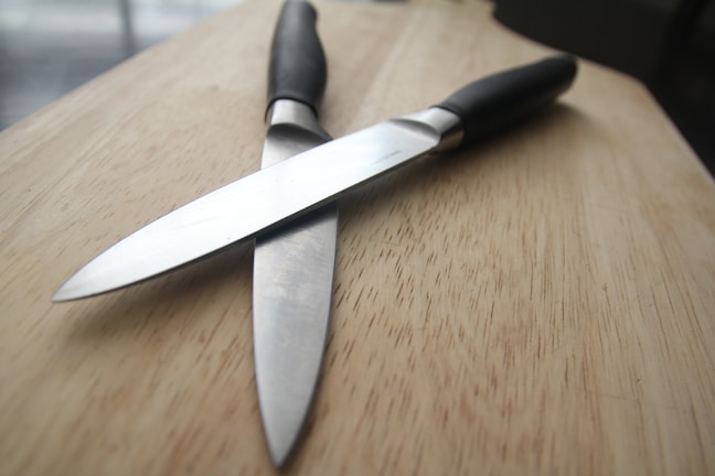 Two kitchen knives with black handles are positioned in a crisscross pattern on a wooden surface. The blades appear to be metallic and reflective, contrasting with the matte texture of the handles. The wooden background is smooth with visible grain patterns, providing a neutral and natural backdrop.