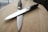 Two kitchen knives with black handles are positioned in a crisscross pattern on a wooden surface. The blades appear to be metallic and reflective, contrasting with the matte texture of the handles. The wooden background is smooth with visible grain patterns, providing a neutral and natural backdrop.