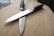 Two kitchen knives with black handles are positioned in a crisscross pattern on a wooden surface. The blades appear to be metallic and reflective, contrasting with the matte texture of the handles. The wooden background is smooth with visible grain patterns, providing a neutral and natural backdrop.