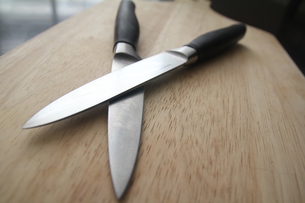 Two kitchen knives with black handles are positioned in a crisscross pattern on a wooden surface. The blades appear to be metallic and reflective, contrasting with the matte texture of the handles. The wooden background is smooth with visible grain patterns, providing a neutral and natural backdrop.
