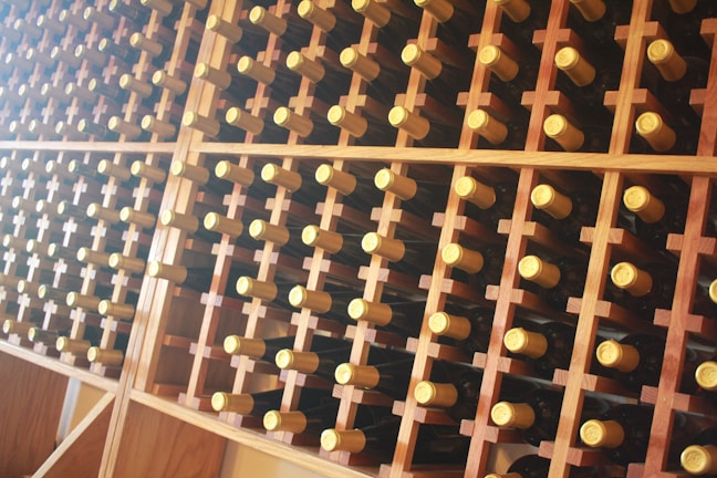 A well-organized wine rack filled with numerous bottles of wine. The bottles are stored horizontally, showcasing their gold foil tops. The rack is made of wooden compartments arranged in a grid pattern.