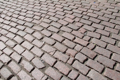 A detailed pattern of cobblestone pavement with uneven, geometric bricks in earthy and muted colors, tightly laid in a slightly curved arrangement. The cobblestones are weathered, with some small gaps filled with dirt and debris.