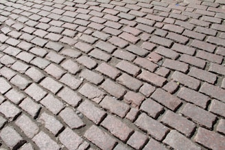 A detailed pattern of cobblestone pavement with uneven, geometric bricks in earthy and muted colors, tightly laid in a slightly curved arrangement. The cobblestones are weathered, with some small gaps filled with dirt and debris.