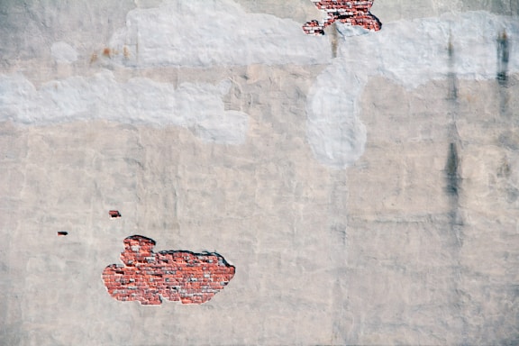 A worn plaster wall with areas of exposed red brick, configured in irregular shapes. Some sections have remnants of painted white clouds atop the plaster, contributing to an abstract artistic feel.