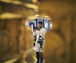 A person in a white shirt is focusing a camera in a formal indoor setting. There is a camera on a tripod in front of them, and the image is framed by blurred golden decor elements.
