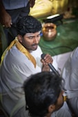 A man dressed in traditional white attire with a golden border is holding a microphone. He has a streak of white paste on his forehead, indicating a cultural or religious mark. In the background, there is a copper vessel placed on a green surface. Another individual is partially visible, also in traditional clothing.