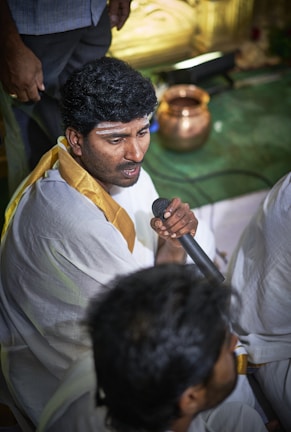 A man dressed in traditional white attire with a golden border is holding a microphone. He has a streak of white paste on his forehead, indicating a cultural or religious mark. In the background, there is a copper vessel placed on a green surface. Another individual is partially visible, also in traditional clothing.