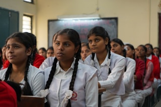 A classroom of engaged students, primarily young women, are sitting in rows wearing white uniforms with braided hair. They appear to be focused and attentive, suggesting a classroom setting. The sunlight filters through the windows, creating a warm ambiance.