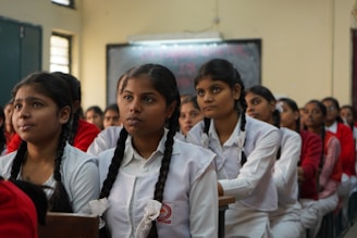 A classroom of engaged students, primarily young women, are sitting in rows wearing white uniforms with braided hair. They appear to be focused and attentive, suggesting a classroom setting. The sunlight filters through the windows, creating a warm ambiance.