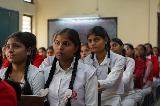 A classroom of engaged students, primarily young women, are sitting in rows wearing white uniforms with braided hair. They appear to be focused and attentive, suggesting a classroom setting. The sunlight filters through the windows, creating a warm ambiance.