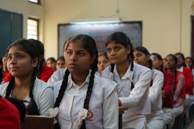 A classroom of engaged students, primarily young women, are sitting in rows wearing white uniforms with braided hair. They appear to be focused and attentive, suggesting a classroom setting. The sunlight filters through the windows, creating a warm ambiance.
