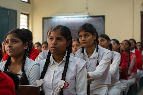 A classroom of engaged students, primarily young women, are sitting in rows wearing white uniforms with braided hair. They appear to be focused and attentive, suggesting a classroom setting. The sunlight filters through the windows, creating a warm ambiance.