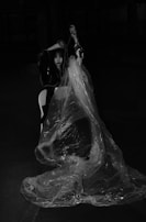 A person is sitting on a chair, partially draped in transparent plastic wrap, in a dimly lit space. The setting appears dramatic, with the play of shadows adding to the mysterious atmosphere.