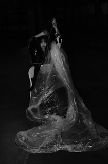 A person is sitting on a chair, partially draped in transparent plastic wrap, in a dimly lit space. The setting appears dramatic, with the play of shadows adding to the mysterious atmosphere.