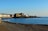 A scenic coastal view featuring a pebble beach in the foreground with a few people relaxing. In the distance, a row of pastel-colored buildings lines the shoreline, alongside a large historic structure. There is a calm sea with reflections and a clear blue sky above.