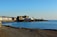 A scenic coastal view featuring a pebble beach in the foreground with a few people relaxing. In the distance, a row of pastel-colored buildings lines the shoreline, alongside a large historic structure. There is a calm sea with reflections and a clear blue sky above.