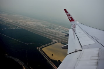 An airplane wing with 'AirAsia' branding is visible, flying over a landscape that includes a large expanse of forest, multiple runways, and what appears to be a water reservoir. The view is slightly hazy, with the airport infrastructure and surrounding greenery seen from a high altitude.