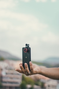 A hand is holding a smartphone with an attached clip-on lens against a blurred background. The phone has a red lens add-on, suggesting a focus on photography or video capture. The sky is overcast, providing a soft light.
