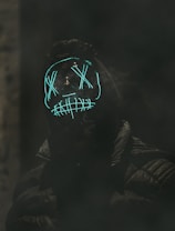 A person is wearing a dark hooded jacket and a glowing mask with neon blue lights forming Xs over the eyes and a stitched pattern over the mouth, creating a mysterious and eerie appearance. The background is dark and smoky, enhancing the unsettling atmosphere.