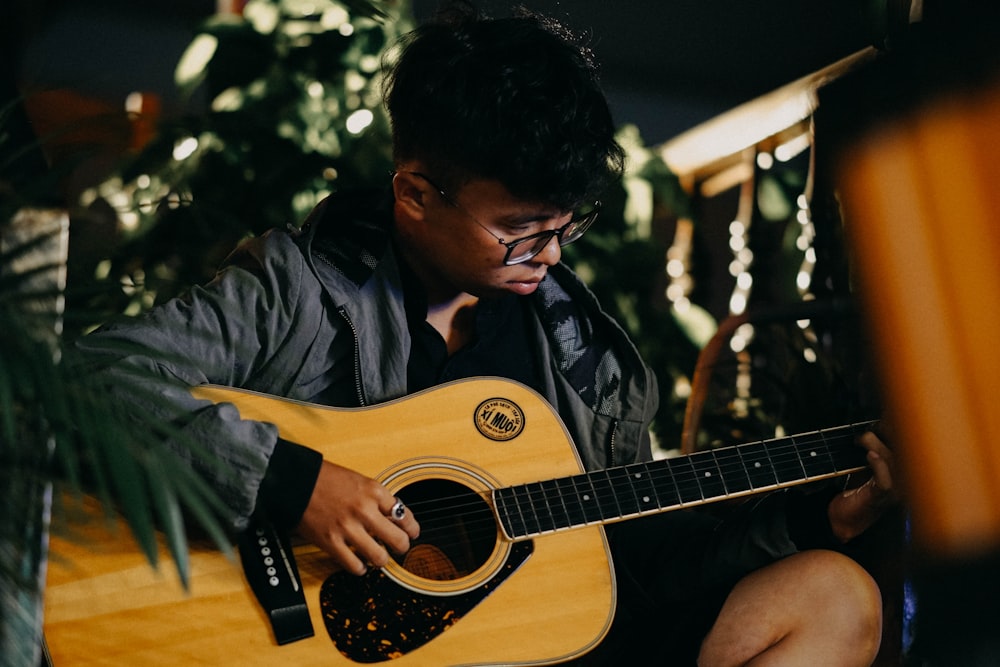 man wearing black jacket and eyeglasses sitting while playing acoustic guitar