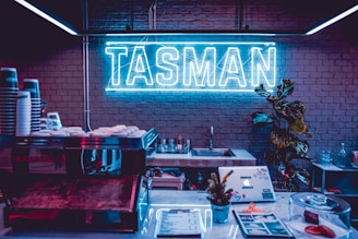 A neon sign reading 'TASMAN' is mounted on a brick wall, illuminating the room with a vibrant blue glow. Below the sign is a countertop with coffee cups stacked on a machine, a menu, and a small potted plant. A sink is positioned behind the counter, and a decorative plant adds greenery to the scene.