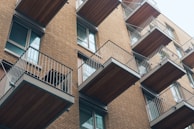 A modern apartment building featuring multiple balconies with metal railings and wooden flooring. The structure is built with light brown bricks and has large windows adjacent to each balcony. The balconies are staggered, creating a visually interesting pattern.