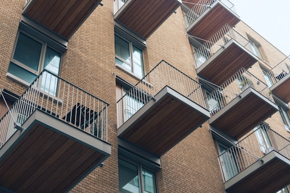 A modern apartment building featuring multiple balconies with metal railings and wooden flooring. The structure is built with light brown bricks and has large windows adjacent to each balcony. The balconies are staggered, creating a visually interesting pattern.