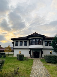 A two-story house with a white facade and dark window frames is surrounded by a neatly maintained garden. A stone path leads up to the entrance, lined with trimmed bushes and trees. The sky above is filled with gray and white clouds, with hints of a setting sun creating a warm glow.