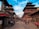 A bustling historic street with traditional Nepalese architecture, featuring tiered pagoda-style buildings. People are walking and sitting along the path, soaking in the cultural atmosphere. The bright sky contrasts with the rich red and brown tones of the buildings.