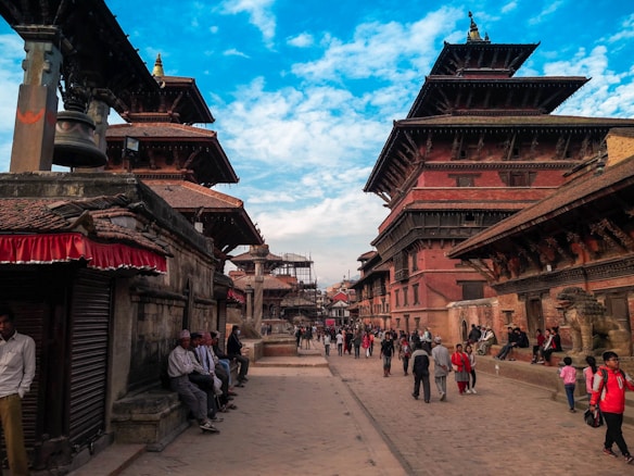 A bustling historic street with traditional Nepalese architecture, featuring tiered pagoda-style buildings. People are walking and sitting along the path, soaking in the cultural atmosphere. The bright sky contrasts with the rich red and brown tones of the buildings.