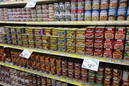 A grocery store shelf filled with various canned goods, featuring prominently several flavors of SPAM and corned beef. The top shelf contains a variety of canned seafood, including clams and tuna, while the lower shelves display canned chili and hash.