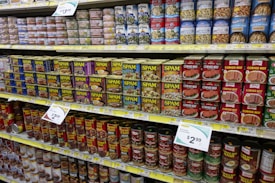 A grocery store shelf filled with various canned goods, featuring prominently several flavors of SPAM and corned beef. The top shelf contains a variety of canned seafood, including clams and tuna, while the lower shelves display canned chili and hash.