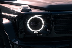 A close-up view of a luxury SUV's front headlight with a distinctive circular LED design. The vehicle has a sleek, glossy black exterior, and the image captures reflections on the surface, highlighting the vehicle's smooth curves and angular features. The headlight is illuminated, creating a sharp contrast against the dark body.