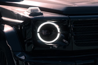 A close-up view of a luxury SUV's front headlight with a distinctive circular LED design. The vehicle has a sleek, glossy black exterior, and the image captures reflections on the surface, highlighting the vehicle's smooth curves and angular features. The headlight is illuminated, creating a sharp contrast against the dark body.