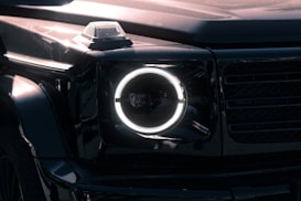 A close-up view of a luxury SUV's front headlight with a distinctive circular LED design. The vehicle has a sleek, glossy black exterior, and the image captures reflections on the surface, highlighting the vehicle's smooth curves and angular features. The headlight is illuminated, creating a sharp contrast against the dark body.