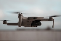 A close-up view of a flying drone with rotating propellers visible. The drone has a sleek, modern design and appears to be lightweight. The background is blurred, giving emphasis to the drone itself.