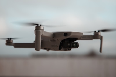 A close-up view of a flying drone with rotating propellers visible. The drone has a sleek, modern design and appears to be lightweight. The background is blurred, giving emphasis to the drone itself.