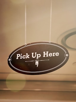 An oval-shaped hanging sign with the text 'Pick Up Here' displayed in bold white letters against a dark background. The sign includes a pointed hand illustration below the text and is framed by a decorative border.
