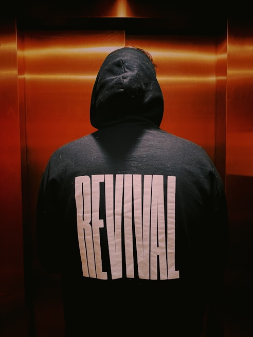 A person stands with their back facing the viewer, wearing a dark hoodie with the word 'REVIVAL' printed in large white letters. They are in an enclosed space with warm, coppery tones on the walls, likely an elevator.